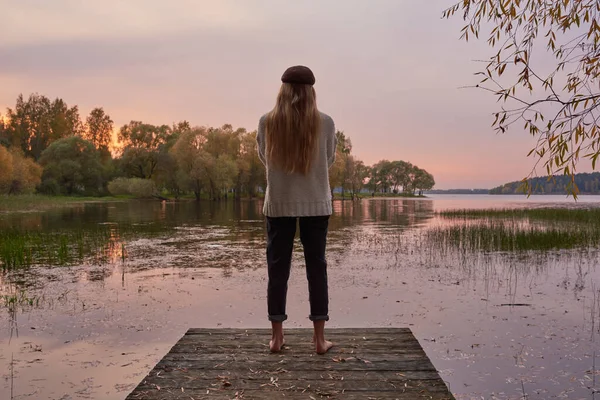Een meisje met lang haar staat op de brug bij het meer met haar rug naar de camera tegen de achtergrond van een zachte zonsondergang. — Stockfoto
