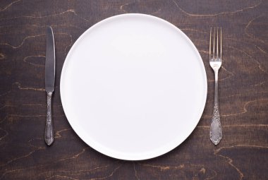 Koyu ahşap masa üzerinde boş beyaz tabak ve gümüş çatal, üst görünümü