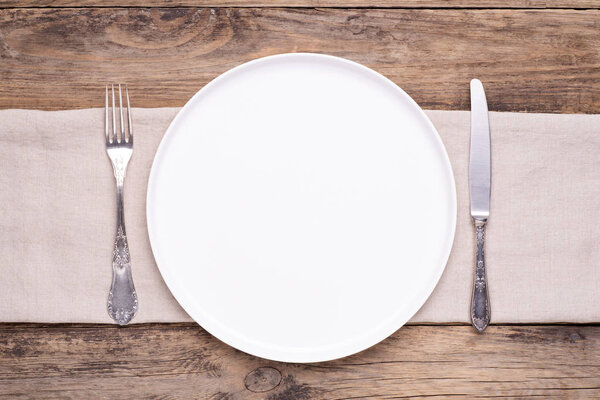 Empty white plate with napkin and silver cutlery on old wooden table, top view