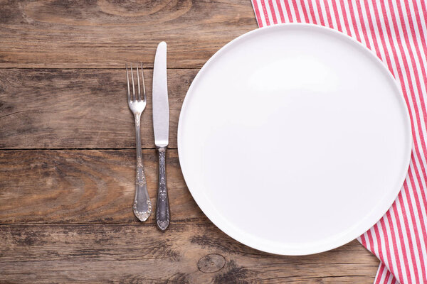 Empty white plate with napkin and silver cutlery on old wooden table, top view with copy space