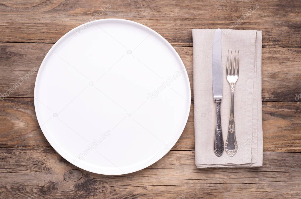 Empty white plate with napkin and silver cutlery on old wooden table, top view