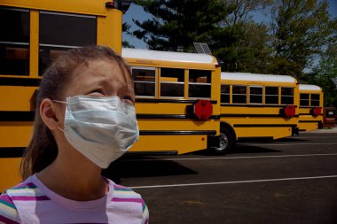 Endişeli genç bir kız okul otobüslerinin önünde duruyor. Toplu taşıma araçlarına binerken onu salgından korumak için maske takıyor.