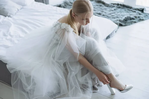 Schöne junge Braut im Kleid zieht Schuhe an, während sie auf dem Bett sitzt — Stockfoto