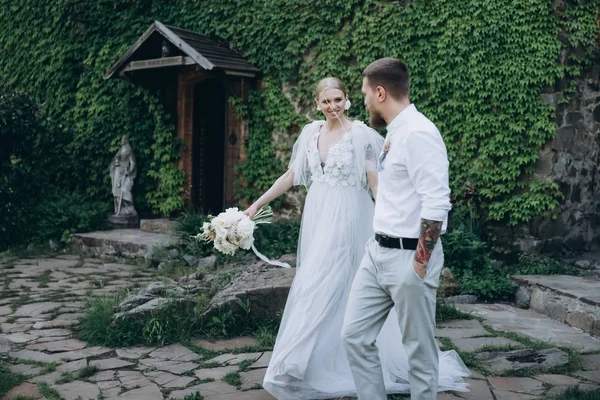 Sposa e sposo passeggiando davanti a un antico edificio coperto di vite e foglie verdi — Foto stock