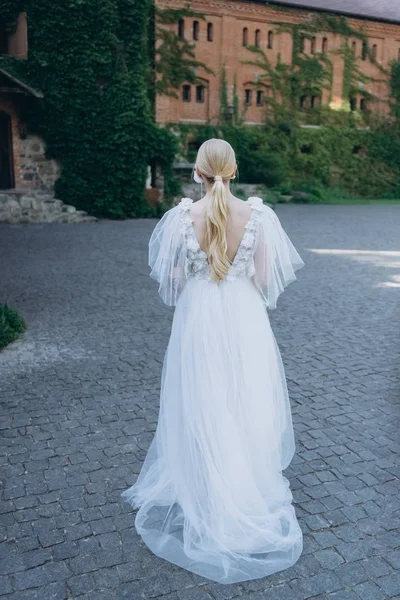 Rear view of young bride in beautiful wedding dress in front of ancient building covered with vine — Stock Photo