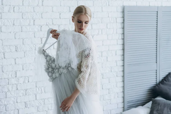 Schöne junge Braut mit Brautkleid auf Kleiderbügel zu Hause — Stockfoto