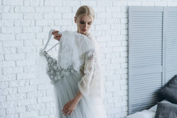 Beautiful young bride with wedding dress on hanger at home — Stock Photo