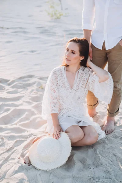 Atractiva mujer con sombrero sentado en la playa y cogido de la mano con el novio - foto de stock