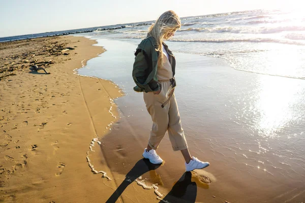 Vista lateral de la mujer joven con ropa elegante caminando en la orilla del mar en el día de verano - foto de stock