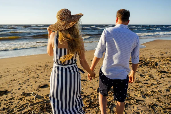 Вид сзади ласковой пары, держащейся за руки и гуляющей по песчаному пляжу в летний день — стоковое фото
