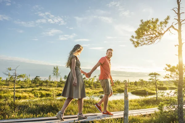 Vista lateral de la pareja cogida de la mano mientras camina sobre un puente de madera con plantas verdes y cielo azul sobre fondo - foto de stock