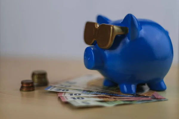 cool blue piggy bank with golden glasses with euro coins and note isolated on wooden table, treasure