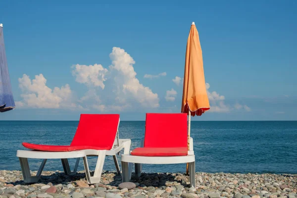 red chaise lounges and orange umbrella on the rocky beach, cloudy sky background