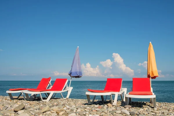 red chaise lounges and orange umbrella on the rocky beach, cloudy sky background