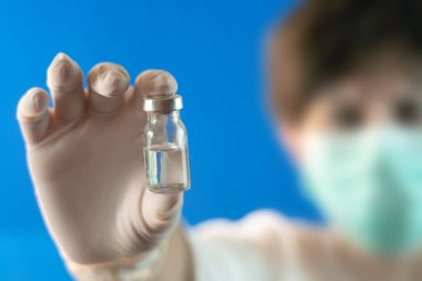 Maskeli ve beyaz eldivenli bir doktor bir ilaç şişesini aşı ile gösteriyor ve seçici bir odak noktası var.