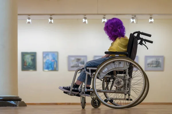 Tekerlekli sandalyede mor saçlı engelli kız sanat galerisini ziyaret ediyor.