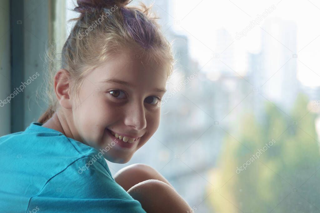 A brown eyed smiling girl in blue t-shirt sitting on the window sill  with blurred outdoor background