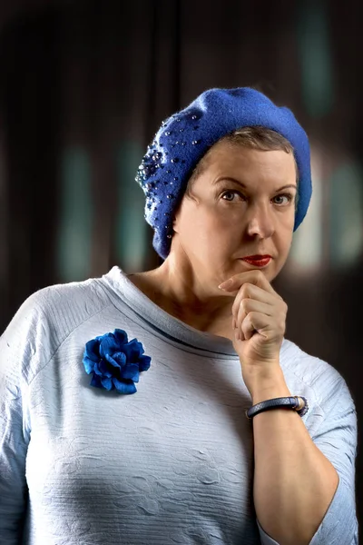 Portrait of the woman of average years in a blue beret, and a blue rose on a breast, and also blue bracelet on a hand. Vertical photo.