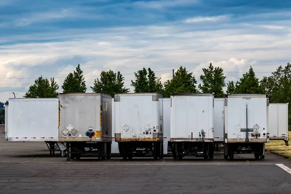 Semi tractor trailers parked in a row with doors closed under a blue cloudy sky with trees