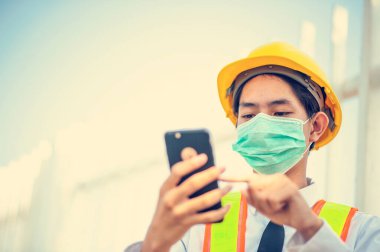 Asyalı mühendis yüz maskesi cep telefonu cihaz teknolojisi inşaat alanında çalışıyor.