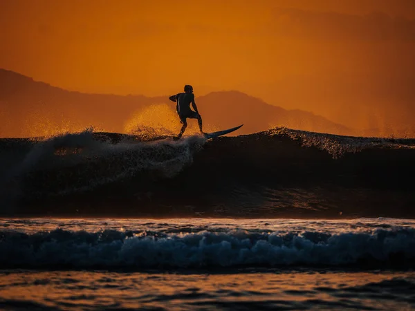 Silhouette of male surfer riding big ocean wave at orange sunset