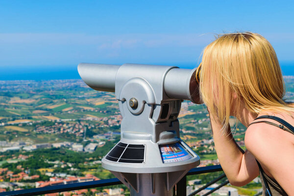 Woman watching beautiful landscape through coin operated telescope in San Marino fortress during sunny and clear summer day