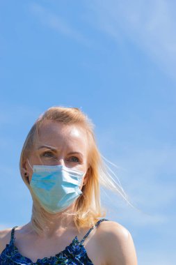 Sağlık ve tıp konsepti. Yüzünde rüzgârlı saçları ve tıp maskesi olan sarışın bir kadının portresi. Kadın mavi gökyüzü arkaplanı ve fotokopi alanı olan mavi elbiseler giyiyor