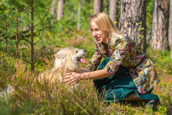 Blonde woman with colorful blouse and green dress playing with beige long haired dog in forest during early autumn time