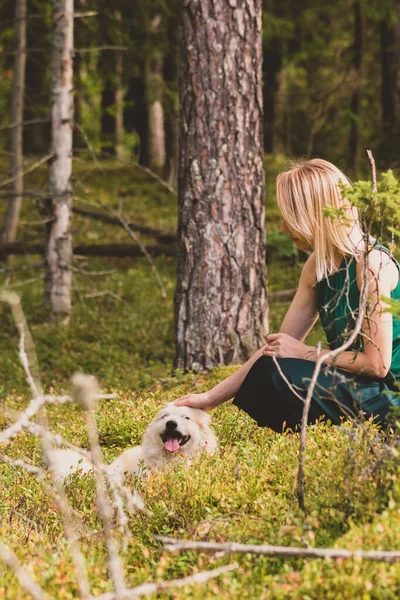 Blonde woman with green blouse and dress playing with beige long haired dog in dense and dark forest during early autumn time
