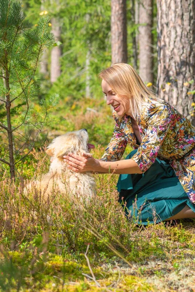 Blonde woman with colorful blouse and green dress playing with beige long haired dog in forest during early autumn time