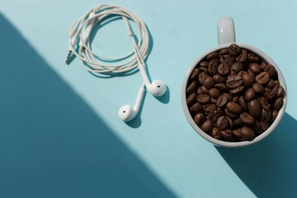 Black coffee beans in blue cup and white headphones on blue background with sunlight shadow top view. Morning drink concept. Cafe and breakfast background. Roasted coffee mug. Aroma drink. Morning relax. Music concept.