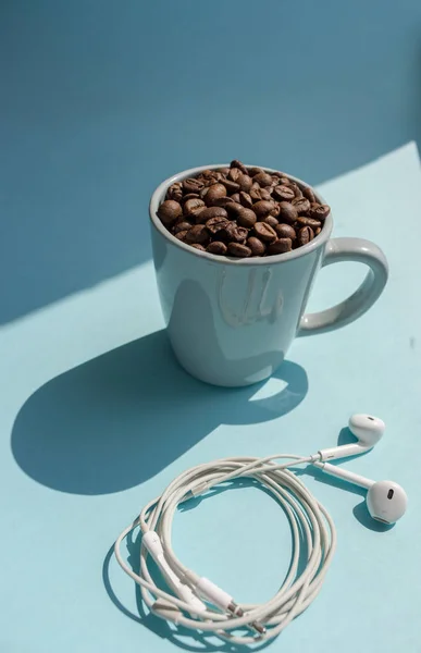 Black coffee beans in blue cup and white headphones on blue background with sunlight shadow. Morning drink concept. Cafe and breakfast background. Roasted coffee mug. Aroma drink. Morning relax. Music concept.