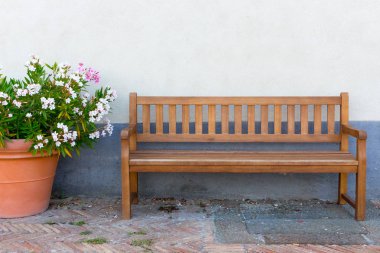 Big clay pot with white and pink flowers and wooden bench against street wall. Outdoor decoration. Gardening design. Patio and relax concept. Large vase with blooming flowers and bench on street. Europe and travel concept.