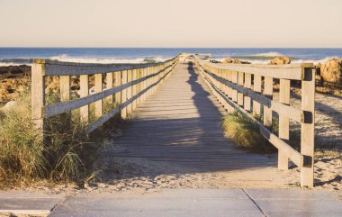 Sahile giden çitli ahşap patika. Sabahları deniz kıyısında yürüyoruz. Seyahat ve yürüyüş konsepti. Camino de Santiago manzarası. Portekiz 'de Atlantik Okyanusu kıyısında. Perspektifte ahşap iskele. 
