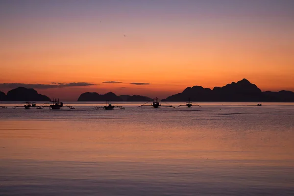 Silhouettes of boats in tropical harbor in the evening. Sunset in lagoon in Philippines, Palawan, El Nido. Sunset on beach. Scenic sunset with mountains isles on horizon. Summer vacation and travel. Exotic landscape.