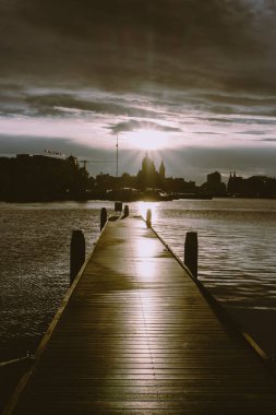 Amsterdam iskelesinde gün batımında tekneler ile ahşap iskele. Akşamları gece karanlığı ışığında Amsterdam şehir siluetleri. Hollanda 'da akşam gökyüzünde katedral silüeti. Amsterdam Rıhtımı. 