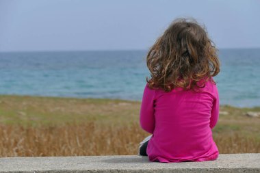 Küçük bir kız alçak bir duvarda oturuyor ve tek başına denize bakıyor.