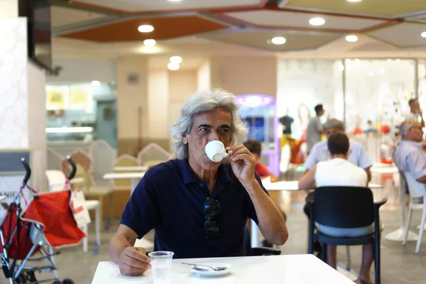 a mature Italian man with long white hair takes a coffee smiling