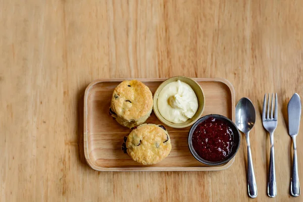 Çilek reçeli, krema ve çatal bıçakla birlikte ahşap bir tepside dinlenen iki çöreğin üst görüntüsü..