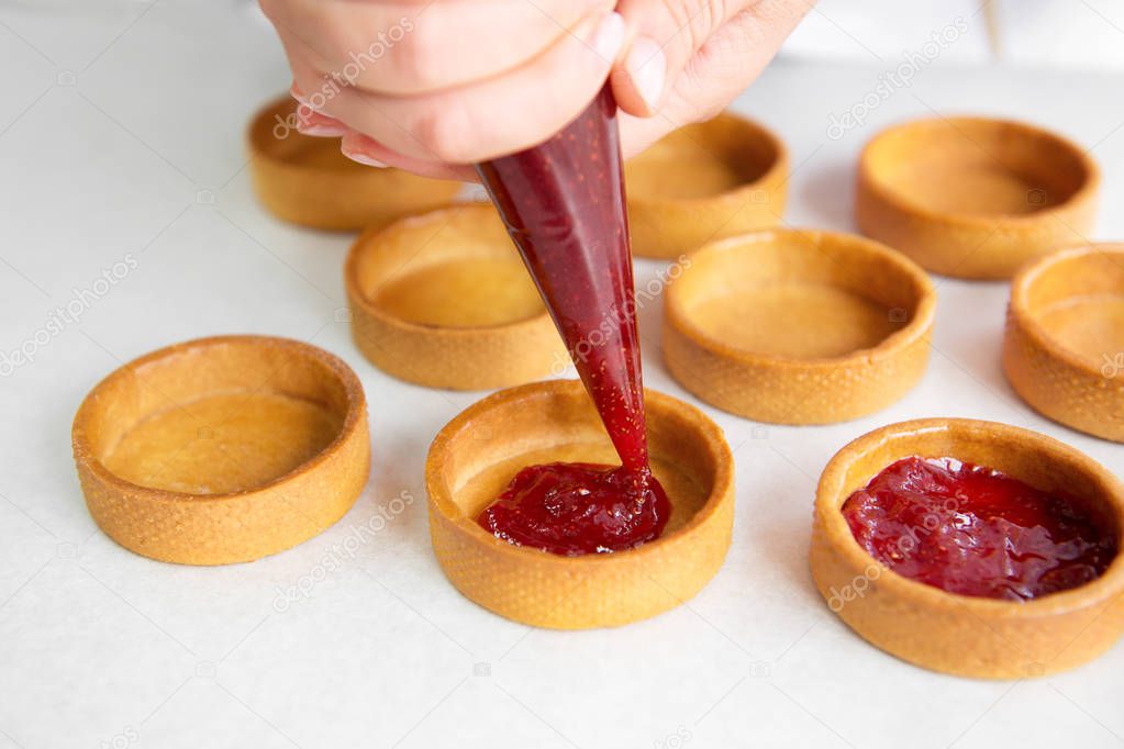 Close-up of pastry chef pouring red filling from piping bag into baked tarts