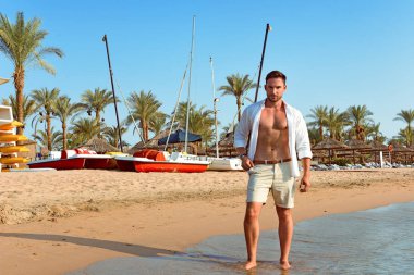 Yakışıklı adam beyaz gömlek daimi bir plajda yaz günü hayattan zevk sıcak bakıyor. Vahşi erkek model su çıplak Göğüs dışarı, sırıtma üzerinde görünüm ve tarz gözleri ile gülümseyen ıslak kısa ile poz veriyor. Tatilde yalnız ve üzgün üzgün yüz adam