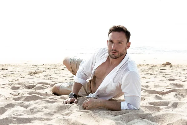 Handsome man in white shirt standing on a beach looking hot on summer day enjoying life. Brutal Male model poses with wet short in water naked chest out, smiling with smirk on his look and kind eyes. Sad face man on vacation alone and depressed