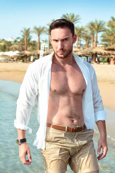 Handsome man in white shirt standing on a beach looking hot on summer day enjoying life. Brutal Male model poses with wet short in water naked chest out, smiling with smirk on his look and kind eyes. Sad face man on vacation alone and depressed