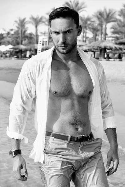 Handsome man in white shirt standing on a beach looking hot on summer day enjoying life. Brutal Male model poses with wet short in water naked chest out, smiling with smirk on his look and kind eyes. Sad face man on vacation alone and depressed