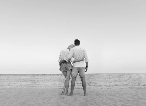 beautiful couple in love enjoying their hawaii vacation looking beach view. Cute blonde woman and handsome bearded man in white shirts standing on a sand hugging, holding hands, watching sea side.