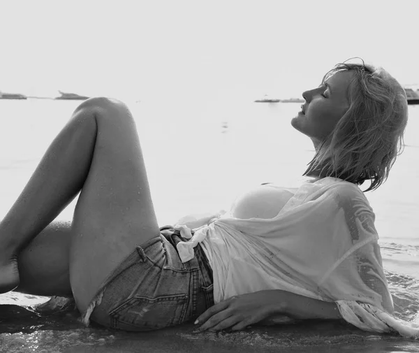 beautiful young blond woman with a sexy body lays down on a sad beach with her clothes wet from sea water on a coast. Blonde model poses with passion on a vacation trip