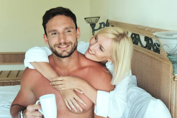 handsome young couple enjoying their morning coffee laying in bed in hotel room happy smiling. Big muscular men and sexy blonde women having fun in bedroom. Family on vacation relaxing laughing huging