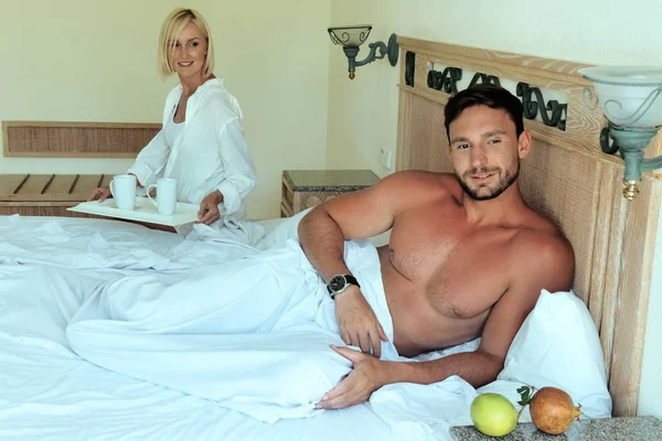 handsome young couple enjoying their morning coffee laying in bed in hotel room happy smiling. Big muscular men and sexy blonde women having fun in bedroom. Family on vacation relaxing laughing huging