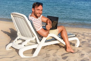 Yakışıklı adam çalışır ve seyahat güzel bir plajda şezlong üzerinde eğleniyor dizlerinin üstünde laptop ile döşeme para kazanmak. Genç milyoner işadamı çiziminden bir kumlu plaj rahatlatıcı üzerinde çalışır