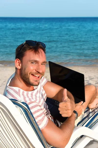 Handsome man works and travels on a beautiful beach laying on chaise lounge with laptop on his knees having fun making money. Young millionaire businessman works his notebook on a sandy beach relaxing
